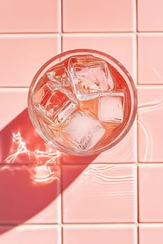 A clear glass filled with ice cubes sits on a pink tiled surface, reflecting soft sunlight, creating a vibrant and refreshing look.