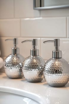 Three elegant silver soap dispensers with a hammered finish sit on a bathroom sink, adding a modern touch to the decor.
