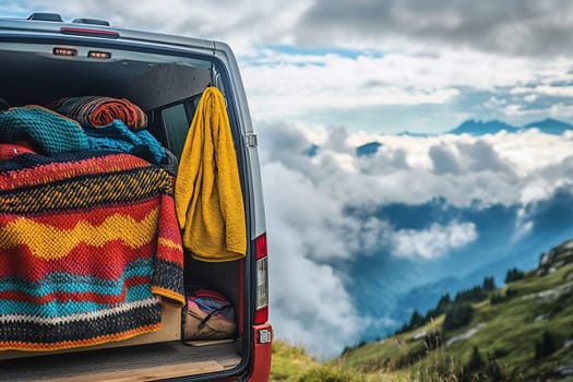 A colorful blanket and shawl hang from a van in a scenic mountain landscape with clouds below and peaks above, showcasing outdoor adventure.