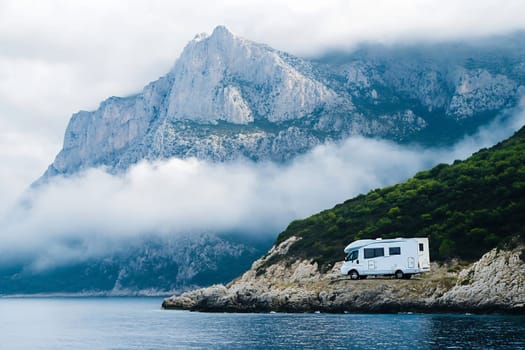 A camper van is parked by the water, surrounded by stunning mountain views and misty clouds on a tranquil coastal day.