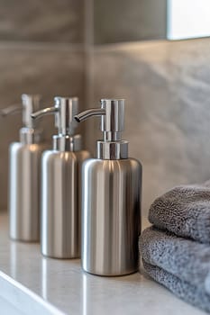 Three sleek stainless steel dispensers sit next to a neatly folded gray towel in a modern bathroom setting. Soft lighting enhances the atmosphere.
