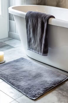 A stylish bathroom features a white bathtub with gray towels draped, complemented by a soft rug and a candle, creating a relaxing atmosphere.