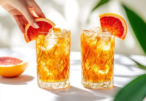 Two tall glasses filled with ice and citrus beverages are placed on a table, garnished with orange slices. A hand adds a slice to one glass.