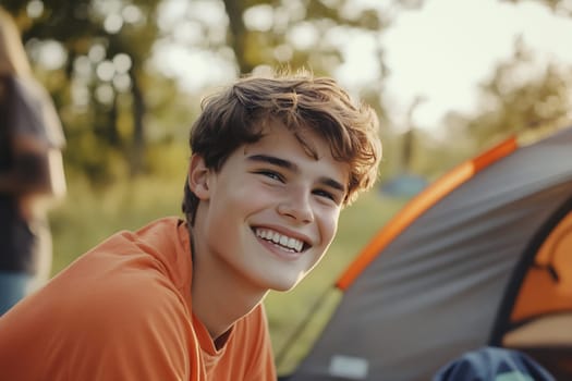 A smiling teenager in an orange shirt relaxes outside a camping tent during a sunny summer evening surrounded by nature.
