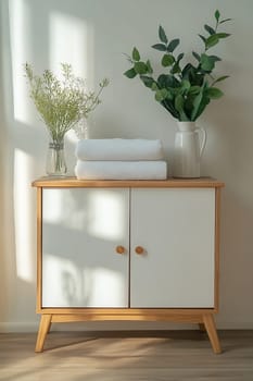 A wooden cabinet with white doors holds fresh towels. Two vases with greenery decorate the top, illuminated by soft sunlight.