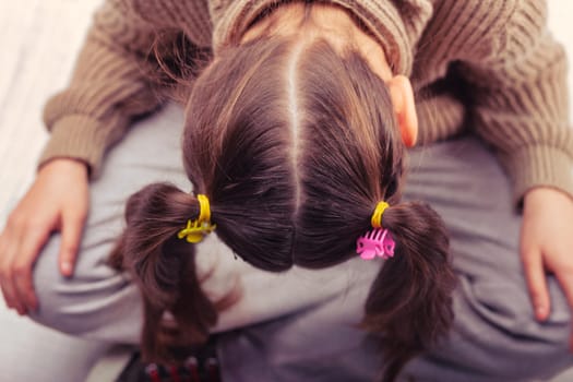 Top view of a young girl with pigtails and colorful hair clips, hiding her face, wearing a knitted sweater and gray trousers, expressing shyness or sadness