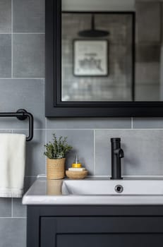 A stylish bathroom features a sleek sink, gray tiles, a decorative plant, and a black mirror enhancing the contemporary atmosphere.