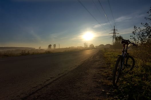 Sunset in the background of the road and fields with grass and trees.