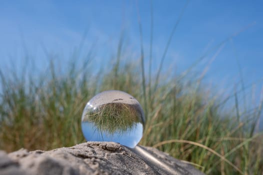 A glass ball lies in the sand of a dune by the sea, the dune grass and the blue sky are reflected in the ball