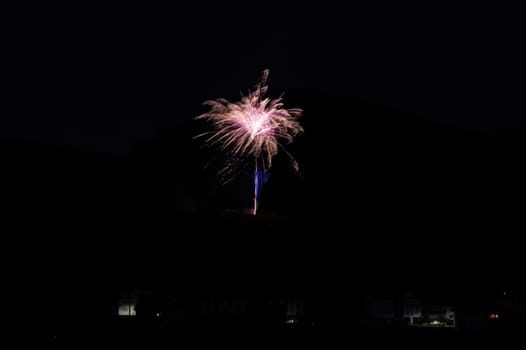 A firework rocket in the sky on a mountain at night, a few illuminated houses in the foreground