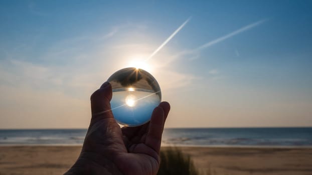 A hand of an  person holds a glass ball, the sea and the setting sun are reflected in the ball