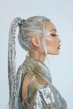 A woman with striking silver hair styled in braids poses elegantly, showcasing a shimmering outfit and bold earrings against a light background.