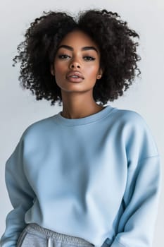 A young woman poses confidently in a light blue sweatshirt, showcasing her natural curls and serene expression against a plain background.