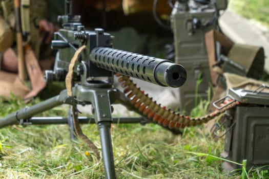 A black machine gun with a long barrel is on a grassy field. The gun is surrounded by a lot of ammunition