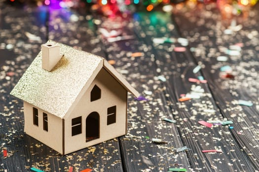 A decorative miniature house stands on a wooden table surrounded by colorful confetti and sparkling lights celebrating the holiday season.
