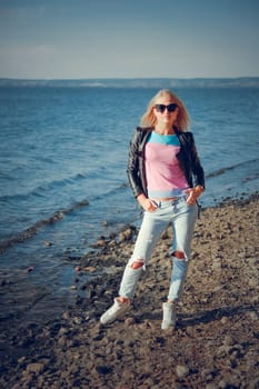Young blond woman wearing sunglasses and a leather jacket posing on a pebbly beach