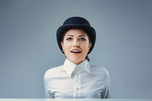 Portrait of a surprised young businesswoman looking up while wearing a bowler hat and a white shirt, isolated on a gray background, expressing amazement and curiosity