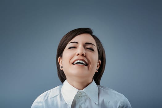 Studio portrait of a businesswoman laughing with her eyes closed, wearing a white shirt against a blue-gray background, expressing joy and happiness