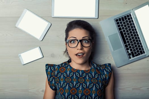 Young adult woman lying on the floor with several electronic devices around her is feeling overwhelmed by technology and information overload
