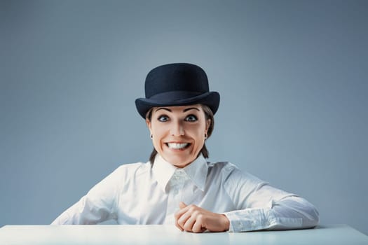 Confident young businesswoman in a bowler hat smiles joyfully at the camera in a quirky office setting with a blue background