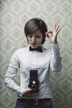 Young female photographer wearing a bow tie and holding a vintage twin-lens reflex camera gestures okay with her hand in front of a patterned wallpaper background