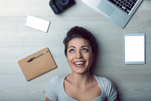 Young woman is lying on a wooden floor, smiling and looking up, surrounded by various technological devices, including a laptop, tablet, smartphone, camera, and notebook