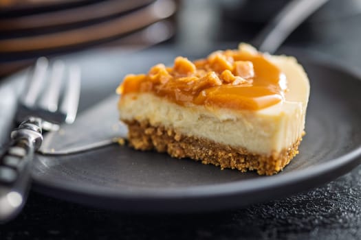 A cut piece of cheesecake with caramel topping on plate on a black table.