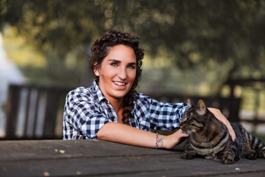 Young woman with curly hair, wearing a checkered shirt, smiles while petting a tabby cat lying on a wooden table in a park-like setting, with trees in the background