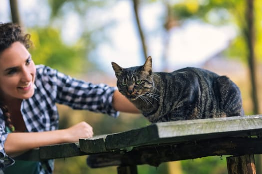 Young woman smiles while reaching out to pet a tabby cat sitting on a wooden table in a garden setting, enjoying a moment of connection with her feline friend