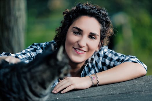Young woman with curly hair, wearing a checkered shirt, smiles at the camera while petting a tabby cat outdoors, resting on a wooden surface