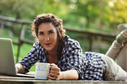 Young woman in her twenties works remotely in a peaceful park, using her laptop and enjoying a hot drink