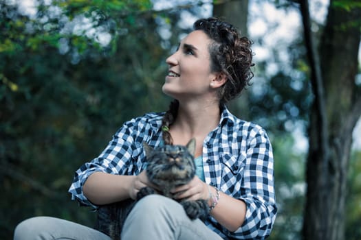 Young woman sits in a forest, holding her tabby cat, looking up with a smile, enjoying nature. They share a moment of love and happiness, surrounded by trees