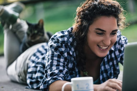 Young woman happily works on her laptop outdoors in a garden with her cat on her back, surrounded by nature and sunlight