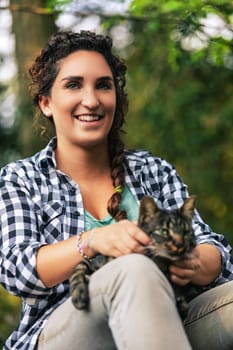 Woman enjoying peaceful moment outdoors with her tabby cat, surrounded by lush greenery. Braided hair, plaid shirt, embodying tranquility. Heartwarming bond between humans and animals