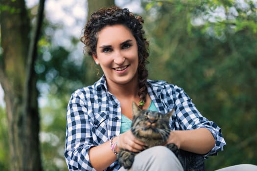 Young woman with curly hair and a checked shirt smiles while holding her tabby cat in a lush forest, enjoying a moment of connection and affection in nature