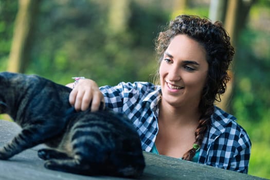Young woman with curly hair, wearing a plaid shirt, smiles as she gently pets a tabby cat on a wooden surface outdoors, creating a heartwarming scene of connection between human and animal