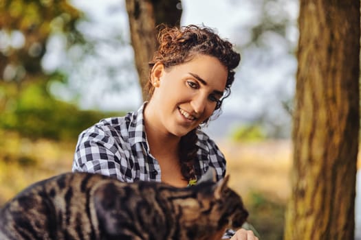 Woman smiling, holding her tabby cat outdoors among trees, radiating love and happiness in the serene nature. The autumn sunlight enhances their bond