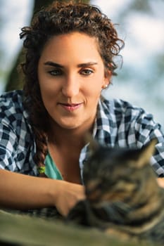 Young woman with curly hair peacefully pets a tabby cat outdoors, basking in warm sunlight and showcasing the bond between humans and animals