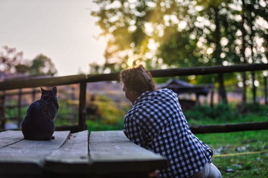 Farmer and her cat are sitting on a wooden table, enjoying the peaceful sunset on a farm, creating a heartwarming scene of companionship and rural tranquility