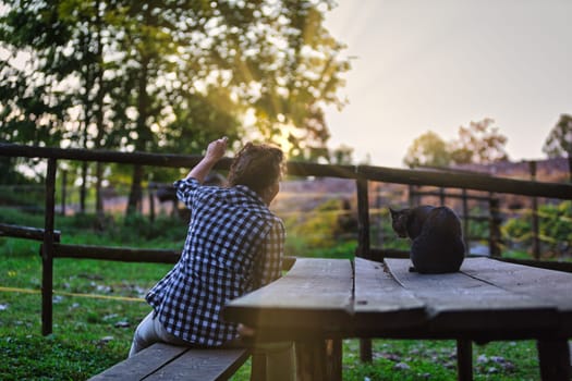 Farmer sits on a wooden bench, gesturing towards animals in a fenced area while a black cat sits on a nearby picnic table, all bathed in the warm glow of the setting sun