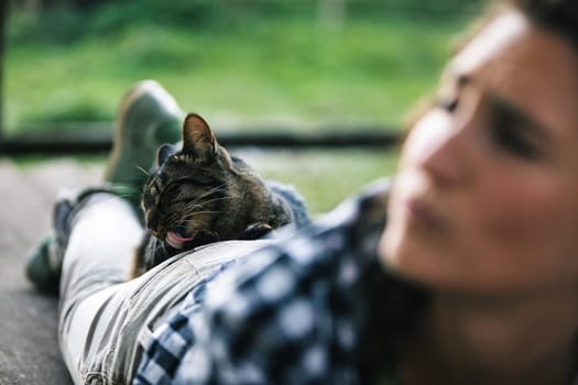 Tabby cat is licking its fur while lying on the back of a farmer woman who is lying on a wooden surface outdoors, enjoying a moment of peace and quiet