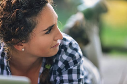 Young woman with curly brown hair, wearing a checkered shirt, lies down and looks away with a pensive expression on her face, in a blurred garden background