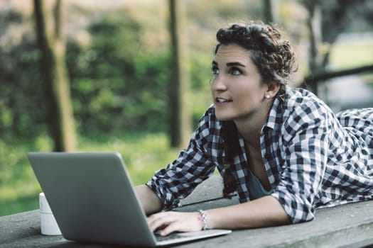 Young woman lying on a table in a park, using a laptop while enjoying the fresh air and natural surroundings, thinking creatively and looking up at the sky