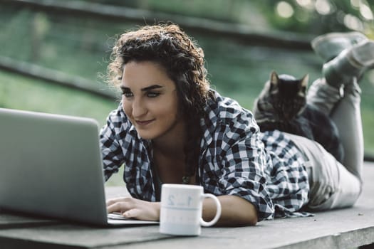 Young woman lying on her stomach on a wooden surface outdoors, working on a laptop while enjoying a mug beside her and a cat peacefully resting on her back