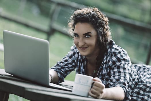 Young woman lying on a wooden table outdoors, working on her laptop while holding a coffee mug, smiling and looking at the camera, enjoying a sunny summer day in nature