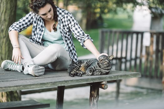 Young woman sitting on a wooden picnic table outdoors gently pets a relaxed tabby cat, enjoying a peaceful moment of connection with the animal