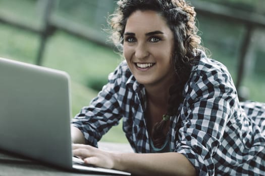 Young woman with curly hair. Wearing a checkered shirt. Lies on a wooden surface outdoors. Diligently working on her laptop. Enjoying the fresh air while embracing modern education and connectivity