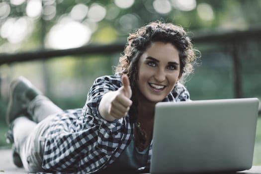 Happy young woman lying in a park, using a laptop while giving a thumbs up, celebrating her success in online education and enjoying the freedom of learning new skills outdoors