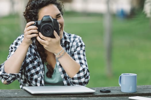 Young adult female photographer smiling while capturing images with a dslr camera in a park, laptop and coffee on the table, embracing her passion for photography