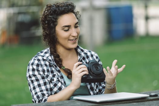 Young adult photography instructor engaging with students during an outdoor workshop, explaining camera settings while utilizing a digital camera and laptop amidst a vibrant park setting